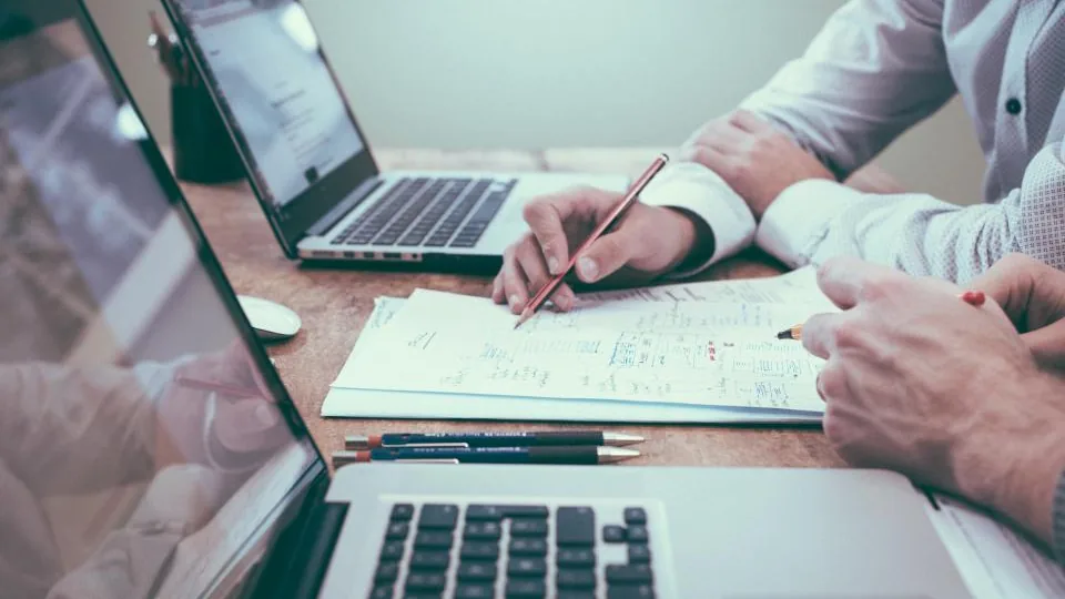 Hands reviewing a printed document with a pen, beside two open laptops