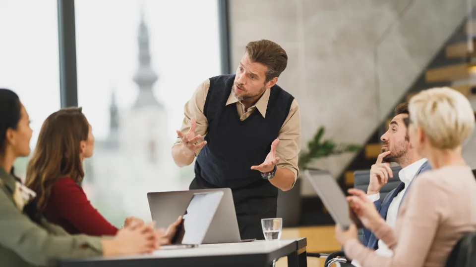 A team meeting around a table with laptops while one colleague stands to present